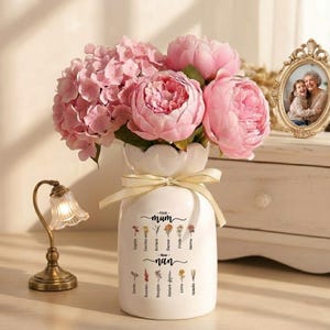 Op de afbeelding: Een witte keramische vaas met een geschulpte bovenkant, vastgebonden met een crèmekleurig lint, bevat roze pioenrozen en hortensia's. De vaas is beschreven met de woorden "First Mum - Name - Nan" en bloemenillustraties. Een kleine lamp en een fotolijst staan op de achtergrond.