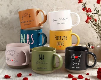Puede incluir: Una colección de tazas de cerámica en varios colores, incluyendo naranja, blanco, azul, amarillo, rosa, verde y negro. Cada taza presenta texto y diseños personalizados, como nombres, fechas y la frase "Forever Love".