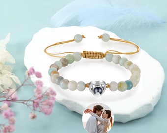 Puede incluir: Un brazalete de cuentas verde claro con un dije de corazón plateado que tiene una foto en su interior. El brazalete tiene un cordón marrón que se puede ajustar para adaptarse a diferentes tamaños de muñeca.