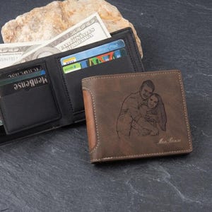 Puede incluir: Cartera de cuero marrón con una imagen grabada personalizada de una pareja. La cartera está abierta, revelando ranuras para tarjetas y efectivo. Otra cartera está al lado. La imagen también muestra efectivo y tarjetas de crédito.