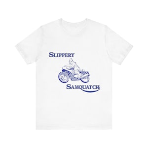 May include: A white short-sleeve t-shirt featuring a blue graphic of a creature riding a motorcycle with the number 16 on the side. The text SLIPPERY is printed above the graphic, and the text SAMQUATCH is printed below the graphic in blue ink.