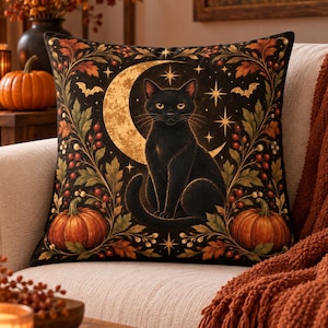 Pode incluir: Uma almofada quadrada com um gato preto sentado em frente a uma lua crescente dourada. O design inclui abóboras cor de laranja, folhas de outono, bagas vermelhas e pequenas estrelas num fundo escuro, emoldurado por padrões botânicos.