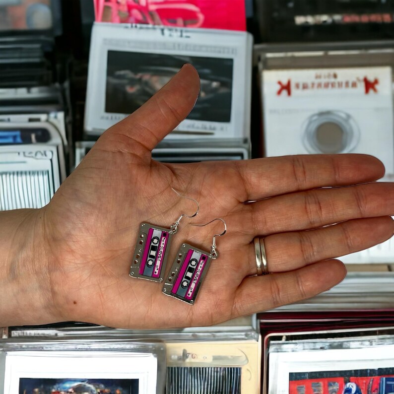 May include: A pair of silver cassette tape earrings with pink and white details. The earrings are hanging from a person's hand.