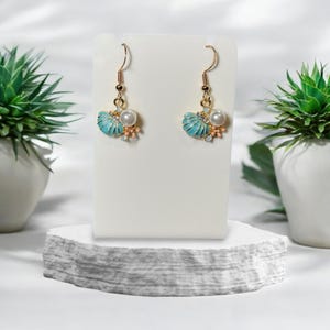 May include: Gold-tone earrings with a seashell design. Each earring has a turquoise shell, a white pearl, and small coral-coloured accents. The earrings are displayed on a white stand.