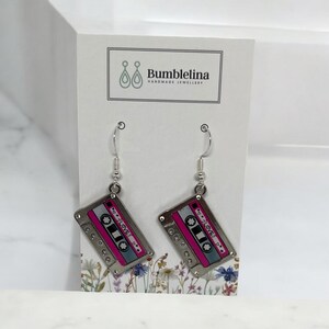 May include: Silver dangle earrings with a pink and silver cassette tape charm. The cassette tape has the word "LOVE" written on it.
