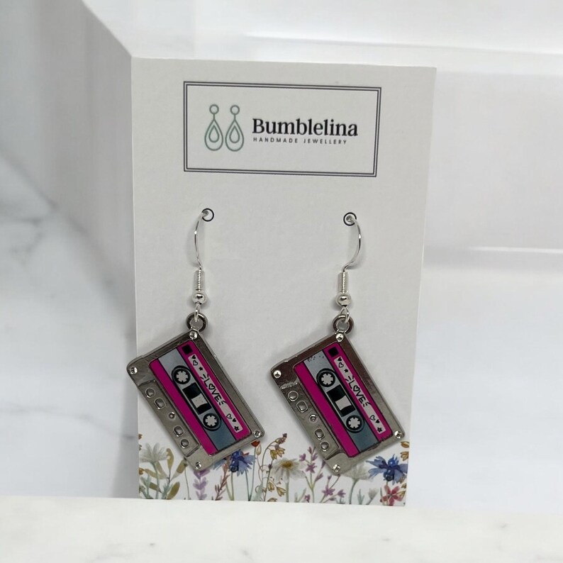 May include: Silver dangle earrings with a pink and silver cassette tape charm. The cassette tape has the word "LOVE" written on it.