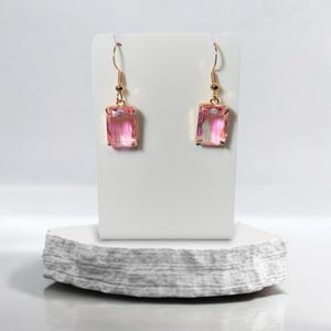 Peut inclure: Une paire de boucles d'oreilles pendantes dorées avec des pierres rectangulaires roses. Les boucles d'oreilles sont présentées sur un support blanc.