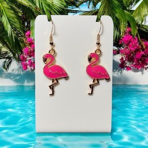 May include: A pair of gold flamingo earrings with pink enamel. The earrings are on a white card with a blue background.