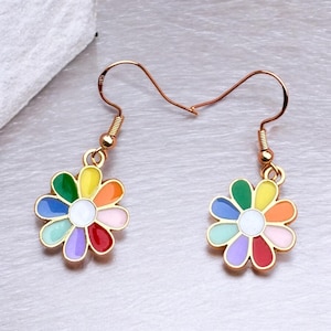 Pode incluir: Um par de brincos dourados com design de margarida. Cada brinco apresenta uma flor esmaltada colorida com pétalas em tons de verde, amarelo, laranja, rosa, vermelho, roxo e azul claro, em torno de um centro branco. Fecho de gancho.