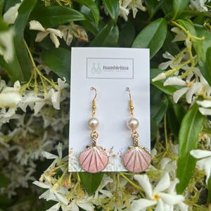 Può includere: Un paio di orecchini dorati con ciondoli a forma di conchiglia rosa e dettagli di perle bianche. Gli orecchini sono esposti su una carta bianca con il testo "Bumblelina Handmade Jewellery".