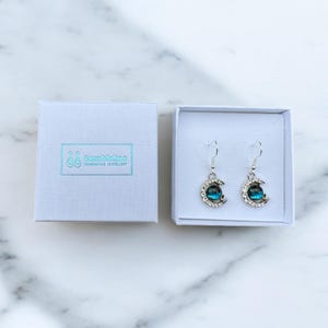Può includere: Un paio di orecchini a forma di mezzaluna d'argento con una pietra preziosa blu al centro di ogni luna. Gli orecchini sono in una scatola bianca con il testo "Buzz Medina Handmade Jewellery".