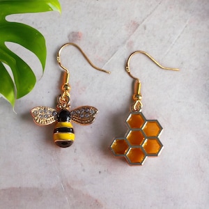 Puede incluir: Un par de pendientes de oro con un diseño de abeja y panal. La abeja es negra y amarilla con brillantes transparentes. El panal es dorado y naranja.