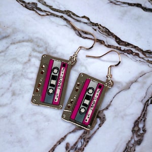 May include: A pair of gold-tone earrings shaped like cassette tapes with a pink and white design. The text "LOVE" is printed on the tapes.