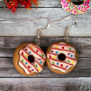 Può includere: Un paio di orecchini a forma di ciambella con glassa rosa e codette colorate. Gli orecchini sono realizzati in pasta polimerica e hanno ganci d'argento.