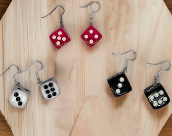 May include: Six pairs of dice earrings in red, black, and white, each with white dots. The earrings are suspended from silver-coloured hooks. The dice are displayed on a light-coloured wooden surface.