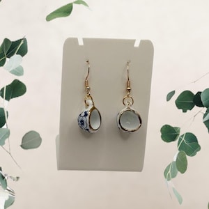 Puede incluir: Un par de pendientes colgantes con mini tazas de té. Las tazas de porcelana blanca tienen diseños florales azules y ribetes dorados. Los pendientes cuelgan de ganchos para la oreja de color dorado.