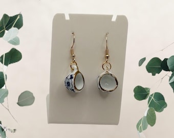 Puede incluir: Un par de pendientes colgantes con mini tazas de té. Las tazas de porcelana blanca tienen diseños florales azules y ribetes dorados. Los pendientes cuelgan de ganchos para la oreja de color dorado.