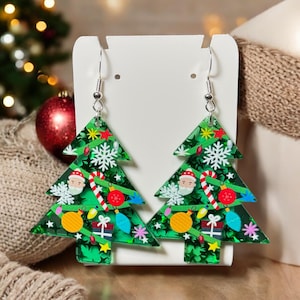 Könnte beinhalten: Ein Paar Ohrringe in Form eines Weihnachtsbaums. Die grünen Ohrringe sind mit festlichen Dekorationen wie Weihnachtsmann-Gesichtern, Schneeflocken, Zuckerstangen und Kugeln verziert. Sie werden auf einer weißen Karte präsentiert.