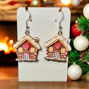 May include: A pair of Christmas-themed earrings. Each earring is shaped like a gingerbread house with a white snow-covered roof, red heart, and striped details. The earrings hang from silver-colored hooks.