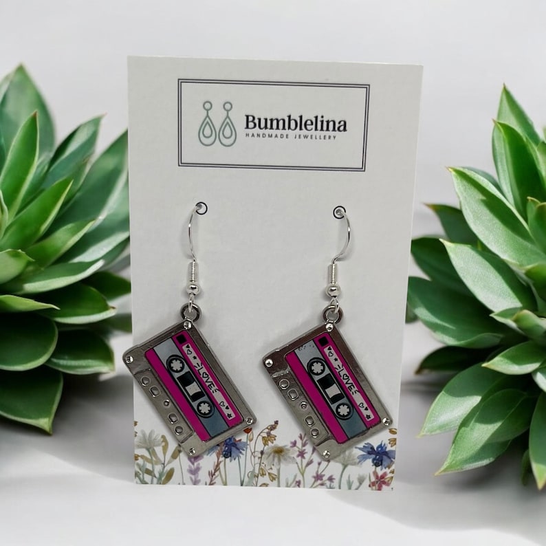 May include: Silver dangle earrings with a pink and silver cassette tape charm. The charm has the text "I &hearts; LOVE" on it.