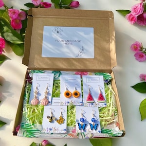 May include: An open cardboard box filled with colorful earrings. The earrings include seashell, sunflower, watermelon, bee, and butterfly designs. The box has a white label that says "YOUR MESSAGE HERE". The box is surrounded by pink flowers and green foliage.