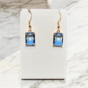 Puede incluir: Un par de pendientes de aro dorados con piedras cuadradas azules. Los pendientes se muestran en una tarjeta blanca.