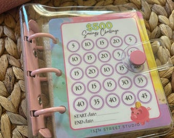 May include: A clear plastic binder with a pink spine and button closure, containing a savings challenge card. The card has a colourful design with circles marked with amounts from £5 to £45, and the text "$500 Savings Challenge". The bottom of the card reads "15TH STREET STUDIO".