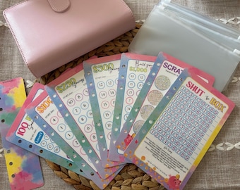 May include: A pink binder and clear plastic pouches are arranged with colourful savings challenge cards. The cards display amounts like £100, £250, and £500, with spaces to mark progress. The items are on a woven surface.