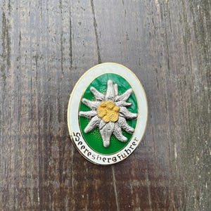 May include: Oval-shaped pin with a green background and a white border. The pin features a silver edelweiss flower with a yellow center. The words "Seeresbergführer" are written in a curved font around the flower.