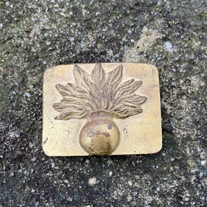 May include: A rectangular brass belt buckle with a detailed floral design. The buckle features a central, rounded element with a stylized flame or flower design above it. The brass has a worn, antique appearance.