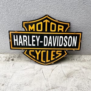 Pode incluir: Uma placa de metal em forma de escudo com o logótipo da Harley-Davidson. O design apresenta um fundo preto com bordas e letras amarelas. O texto diz MOTOR no topo, HARLEY-DAVIDSON no centro e CYCLES na parte inferior.