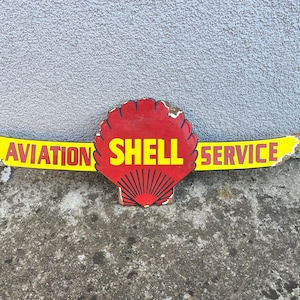 May include: A vintage Shell Aviation Service sign. The sign features a red shell logo with yellow text that reads "SHELL" and a yellow banner with "AVIATION SERVICE" in red text. The sign has a weathered appearance.