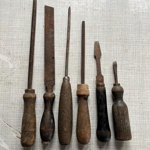 Op de afbeelding: Een verzameling van zes vintage metalen gereedschappen met houten handvatten. De gereedschappen omvatten vijlen en schroevendraaiers, die tekenen van ouderdom en gebruik vertonen. De handvatten variëren in vorm en kleur, van licht- tot donkerbruin en zwart.