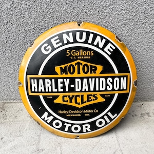 Pode incluir: Uma placa redonda de metal amarela e preta para óleo de motor Harley-Davidson. O texto diz GENUINE, 5 Gallons U.S. MEASURE, MOTOR HARLEY-DAVIDSON CYCLES, TRADE MARK e Harley-Davidson Motor Co MILWAUKEE. WIS. Apresenta ferrugem visível nas bordas.