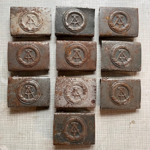 May include: Nine weathered metal belt buckles are displayed. Each buckle has a circular emblem with a central design, encircled by a wreath. The rectangular buckles show signs of age and wear, with a rusty appearance, suggesting they are vintage items.
