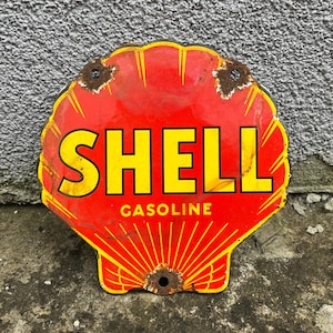 Puede incluir: Letrero de gasolina Shell vintage. El letrero tiene forma de concha, con un fondo rojo y texto amarillo que dice "SHELL GASOLINE". El letrero tiene un aspecto desgastado con manchas de óxido y un aspecto envejecido, lo que sugiere que es antiguo.