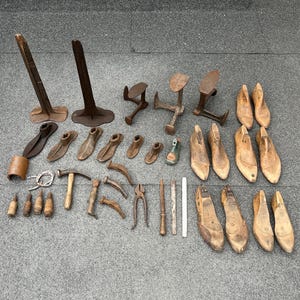 May include: A collection of antique shoemaking tools, including wooden shoe lasts, hammers, pliers, and metal forms. The tools are arranged on a gray surface, showcasing the variety of shapes and sizes used in the craft.