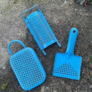 Könnte beinhalten: Drei blaue Küchenutensilien im Vintage-Stil: eine rechteckige Reibe mit Griff, ein rechteckiges Sieb mit Griff und ein kleiner Schöpfer mit Griff. Alle drei Werkzeuge haben mehrere kleine Löcher.