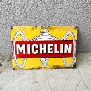 Könnte beinhalten: Ein rechteckiges Metallschild mit gelbem Hintergrund zeigt das Michelin-Maskottchen, eine weiße, aus Reifen bestehende Figur, die eine Zigarre raucht und zwei große weiße Reifen hält. Ein roter Querstreifen in der Mitte zeigt den weißen Text MICHELIN.