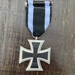 Puede incluir: Medalla de la Cruz de Hierro en negro y plata, con una cinta a rayas azul claro y negro. La medalla presenta una corona, la letra "W" y el año "1870". Un alfiler plateado sujeta la medalla a la cinta.