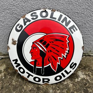 Puede incluir: Letrero redondo vintage de Gasolina y Aceites de Motor con un gráfico de jefe nativo americano. El letrero tiene un borde blanco, detalles en negro y rojo, y las palabras "GASOLINE" y "MOTOR OILS". El letrero muestra signos de edad.