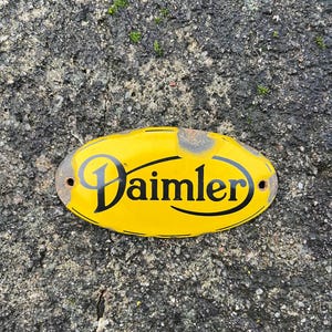 Può includere: Cartello ovale in metallo giallo con la scritta "Daimler" in nero. Il cartello presenta segni di usura e due fori di montaggio. Lo sfondo è una superficie ruvida e grigia.