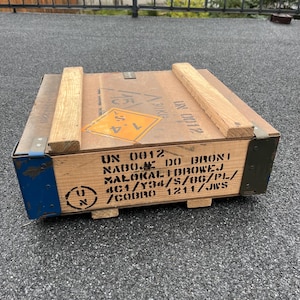 May include: A rectangular wooden crate, light brown in colour with visible wood grain. It features a yellow and orange hazard symbol and black text. The corners are reinforced with blue and green metal. Wooden slats are on top of the crate.