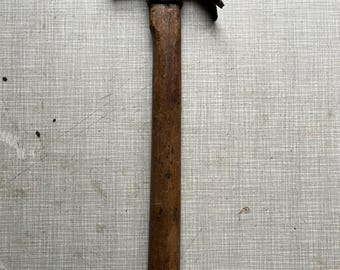 May include: A well-worn claw hammer with a wooden handle and a rusty, metal head. The hammer is positioned vertically against a textured, light gray surface. The claw is slightly curved, and the handle shows signs of use.