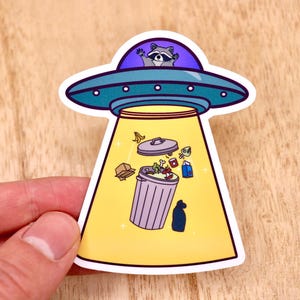 May include: A sticker of a raccoon in a flying saucer, beaming down a trash can full of rubbish. The trash can is purple and the saucer is blue and yellow.