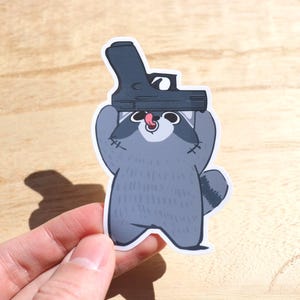 May include: A grey raccoon wearing a black gun on its head. The raccoon is smiling and has a pink tongue sticking out.