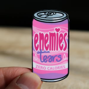 Può includere: Lattina rosa e viola con il testo "enemies tears" e "zero calories" in bianco. La lattina ha un cuore e gocce di pioggia.