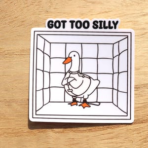May include: A white sticker featuring a cartoon goose with an orange beak and feet, inside a black and white cage. The text "GOT TOO SILLY" is above the cage in black letters.
