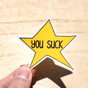 Puede incluir: Una pegatina amarilla en forma de estrella con un borde blanco y las palabras "you suck" en negro. La pegatina está sujeta por una mano sobre un fondo de vetas de madera clara.