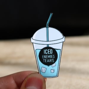 Può includere: Uno spillo in smalto blu e bianco a forma di tazza di tè freddo con una cannuccia. La tazza contiene cubetti di ghiaccio e il testo "ICED ENEMIES TEARS" su un cerchio nero.